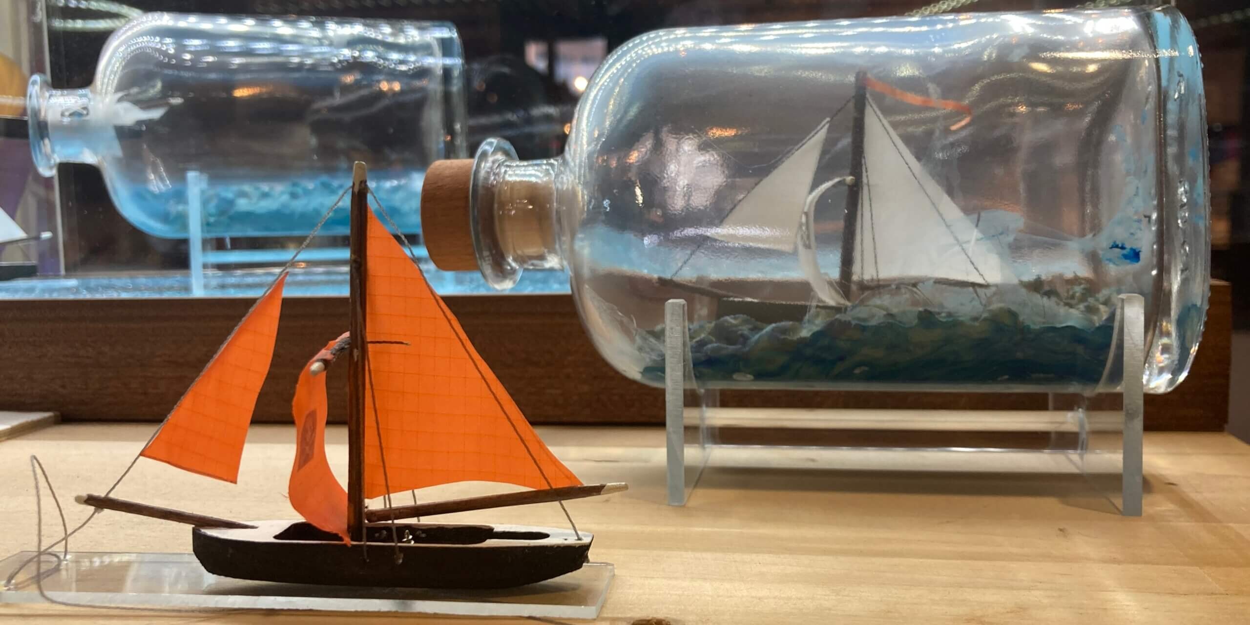 Kleines Holzsegelboot mit leuchtend orangefarbenen Segeln im Vordergrund; rechts daneben ein detailliertes Schiff in einer liegenden Glasflasche auf einem St&auml;nder, beides auf einer hellen Holzplatte vor reflektierenden Lichtern im Hintergrund.