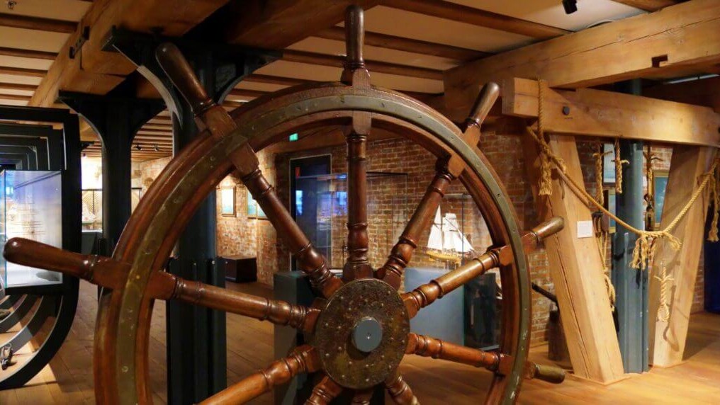 Großes hölzernes Schiffssteuerrad im Vordergrund eines Museumsausstellungsraums mit sichtbaren Holzträgern, Ziegelwand, einem Schiffsmodell im Hintergrund und aufgehängten Tauelementen.