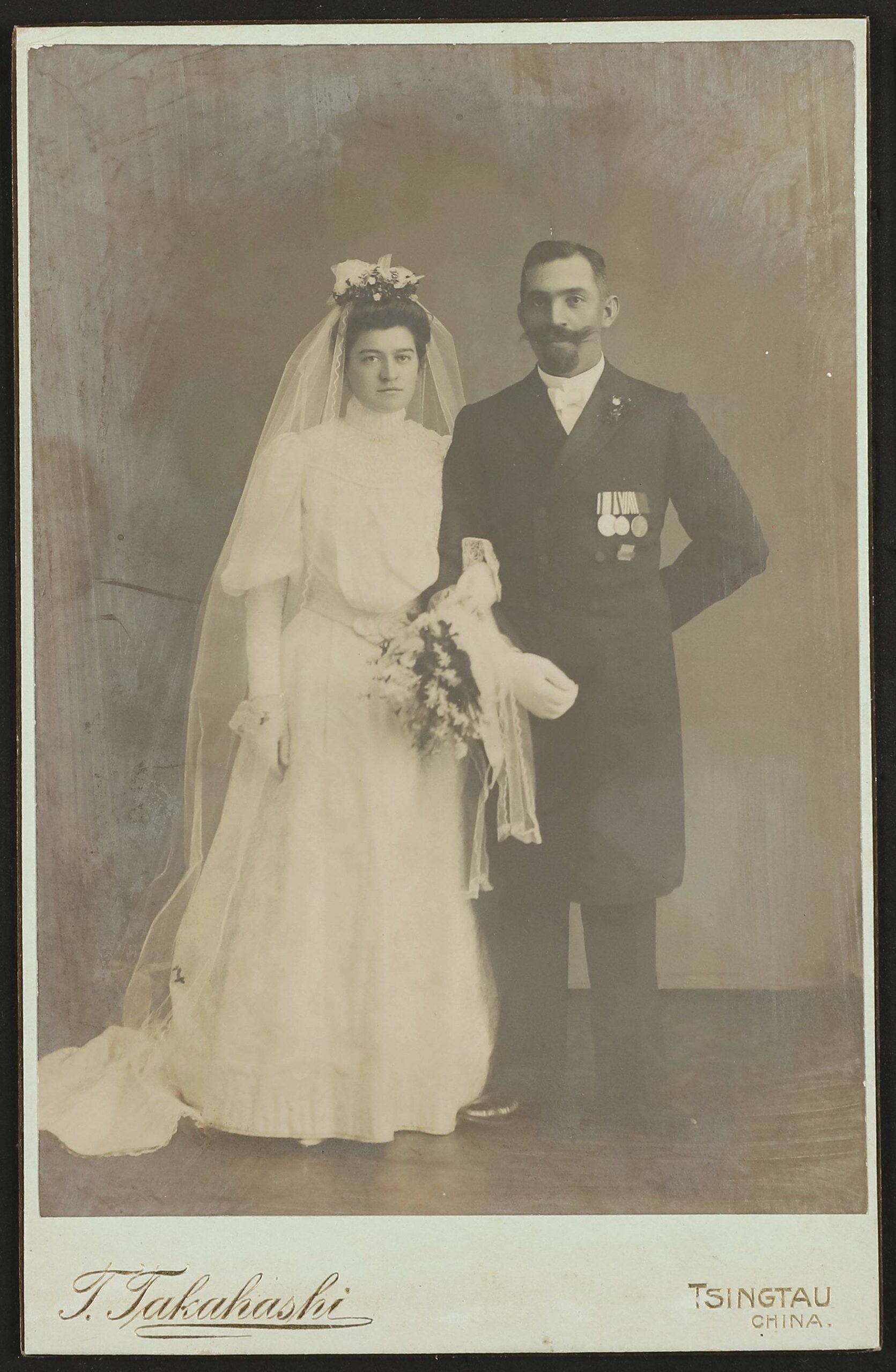 Portr&auml;t eines Brautpaars: Frau in wei&szlig;em Kleid mit Schleier und Strau&szlig;, Mann mit Schnurrbart, Anzug und Medaillen; Aufdruck T. Takahashi Tsingtau