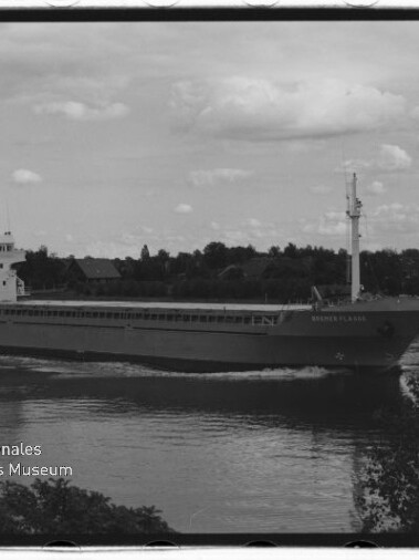 Schwarzweißfoto eines Frachtschiffs auf ruhigem Fluss vor baumbewachsenem Ufer; Wasserzeichen Maritimes Museum Hamburg