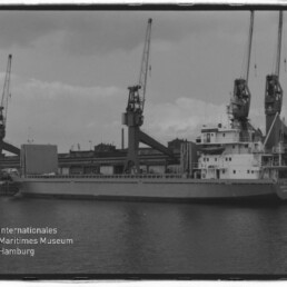 Frachtschiff am Kai mit mehreren großen Hafenkränen im Hintergrund, schwarzweiße Aufnahme mit Museum-Logo unten links.