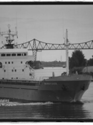 Frachtschiff fährt einen Kanal unter einer Stahlfachwerkbrücke, Uferbebauung im Hintergrund und Museumswasserzeichen unten links.