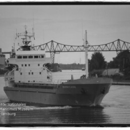 Frachtschiff fährt einen Kanal unter einer Stahlfachwerkbrücke, Uferbebauung im Hintergrund und Museumswasserzeichen unten links.