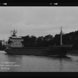 Schwarzweißaufnahme eines Frachtschiffs mit Kommandobrücke am Heck und Mast vor bewaldetem Ufer; Museumssignet unten links.