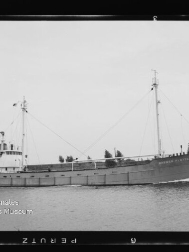 Frachtschiff mit zwei Masten und achterlicher Brücke fährt auf ruhigem Kanal vor baumbesetztem Ufer; Museumslogo unten links.