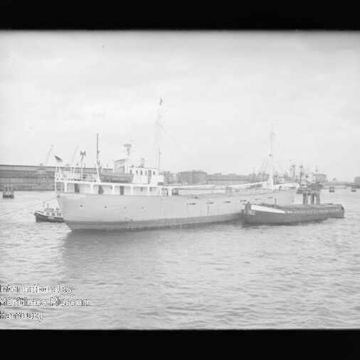Schwarzweißaufnahme eines großen Frachtschiffs mit Schlepper an seiner Seite im Hafen vor Kais und Kränen; Museumslogo unten links
