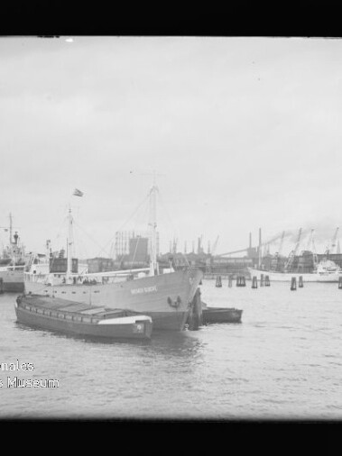 Frachtschiff mit Beiboot im Hafen vor Kaimauern und Kränen; links unten Wasserzeichen 