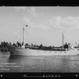 Frachtschiff mit zwei Masten und achterlicher Aufbauten fährt auf ruhigem Fluss, Ufer mit Baumreihe im Hintergrund.