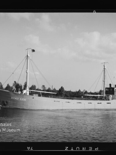 Frachtschiff mit zwei Masten und achterlicher Aufbauten fährt auf ruhigem Fluss, Ufer mit Baumreihe im Hintergrund.