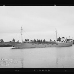Schwarzweißfoto eines Frachtschiffs auf ruhigem Kanal vor Ufer mit Bäumen; unten links Wasserzeichen Maritimes Museum Hamburg.