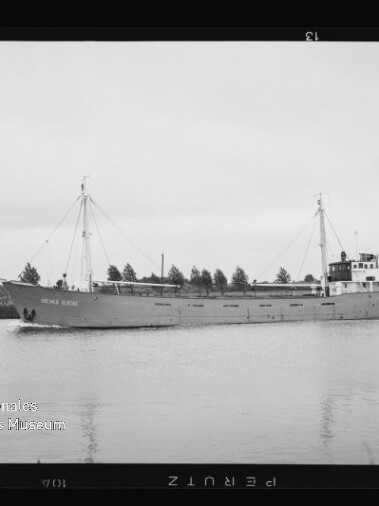 Schwarzweißfoto eines Frachtschiffs auf ruhigem Kanal vor Ufer mit Bäumen; unten links Wasserzeichen Maritimes Museum Hamburg.
