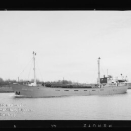 Schwarzweißaufnahme eines Frachtschiffs mit zwei Masten und Brücke, das ruhig auf einem Kanal vor flachem Ufer fährt.