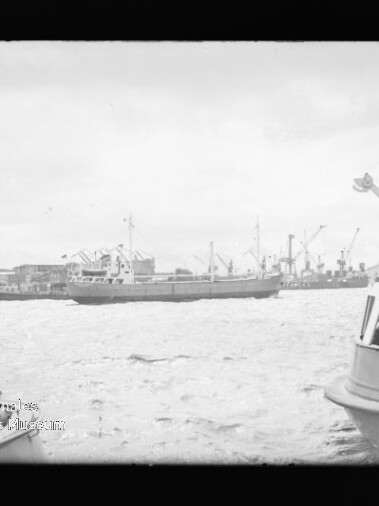 Frachtschiff im Hafen vor Kränen und Lagerhallen, vorn links und rechts Ausflugsschiffe mit Personen und welligem Wasser