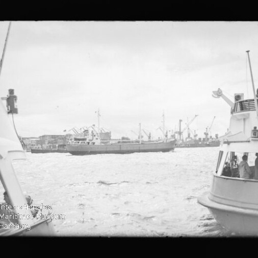 Frachtschiff im Hafen vor Kränen und Lagerhallen, vorn links und rechts Ausflugsschiffe mit Personen und welligem Wasser