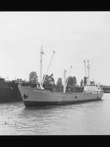 Schwarzweißfoto eines Frachtschiffs mit Ladekränen am Kai, ruhigem Wasser und Museumswasserzeichen unten links