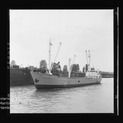 Schwarzweißfoto eines Frachtschiffs mit Ladekränen am Kai, ruhigem Wasser und Museumswasserzeichen unten links