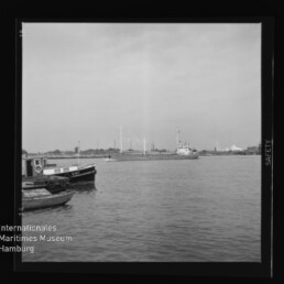 Hafenblick mit kleinen Booten vorn, Frachtschiff und Industrie mit Schornsteinen am anderen Ufer; Museum-Logo unten links.