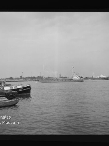 Hafenblick mit kleinen Booten vorn, Frachtschiff und Industrie mit Schornsteinen am anderen Ufer; Museum-Logo unten links.