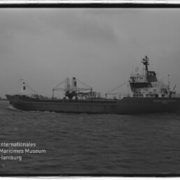 Schwarzweißfoto eines Frachtschiffs mit Ladekranen auf ruhiger See vor Ufer; Museumslogo unten links.