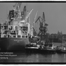 Schwarzweißfoto eines Frachtschiffs im Industriehafen mit Kränen und Schleppern; Wasserzeichen Maritimes Museum Hamburg.