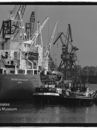 Schwarzweißfoto eines Frachtschiffs im Industriehafen mit Kränen und Schleppern; Wasserzeichen Maritimes Museum Hamburg.