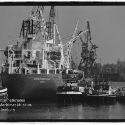 Großes Frachtschiff am Kai, flankiert von Schleppern und Hafenkränen; schwarz-weiß Aufnahme mit Museumslogo.