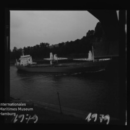 Schwarzweißaufnahme eines Frachtschiffs mit Ladekränen auf einem schmalen Flusskanal, baumbewachsene Ufer im Hintergrund.