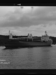 Schwarzweißfoto eines Frachtschiffs mit Ladekränen vor bewaldeter Küste; Text: Internationales Maritimes Museum