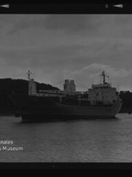 Schwarzweißaufnahme eines großen Frachtschiffs auf ruhigem Wasser vor bewaldeter Küste, mit Filmrand und Wasserzeichen Internationales Maritimes Museum Hamburg.