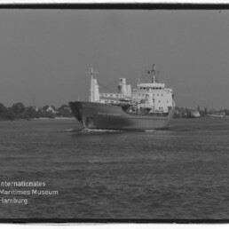 Frachtschiff fährt über ruhige See vor Ufer mit Häusern; linkes Eck zeigt Wasserzeichen des Maritimen Museums Hamburg.