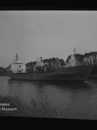 Frachtschiff auf ruhigem Kanal vor einer Baumreihe; links unten Logo und Schriftzug Internationales Maritimes Museum Hamburg.