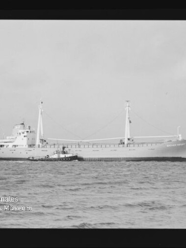 Schwarzweißaufnahme eines Frachtschiffs mit kleinem Schlepper an seiner Seite auf ruhiger See; Museumslogo unten links.