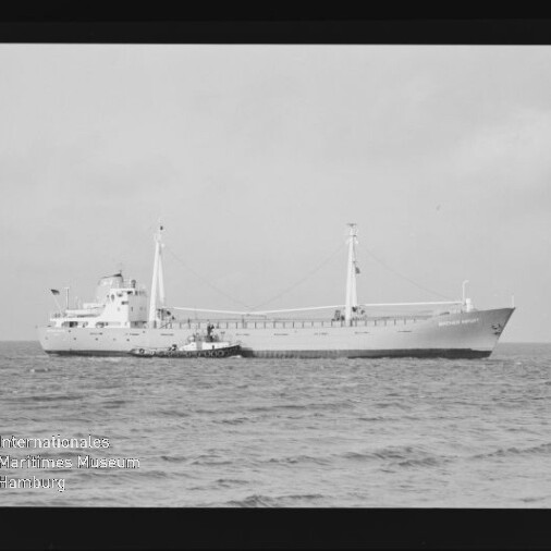 Schwarzweißaufnahme eines Frachtschiffs mit kleinem Schlepper an seiner Seite auf ruhiger See; Museumslogo unten links.
