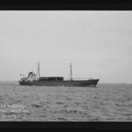 Frachtschiff in Seitenansicht mit Mast und Heckaufbau auf ruhiger See; links unten Museum-Logo.