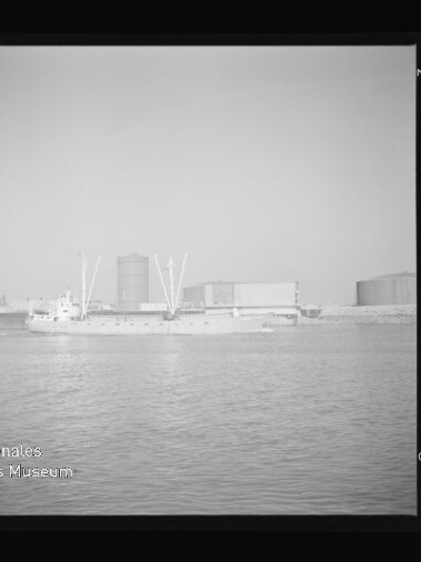 Schwarzweißaufnahme eines Frachtschiffs vor einem Industriehafen mit Tanks und Kränen, Wasser im Vordergrund.