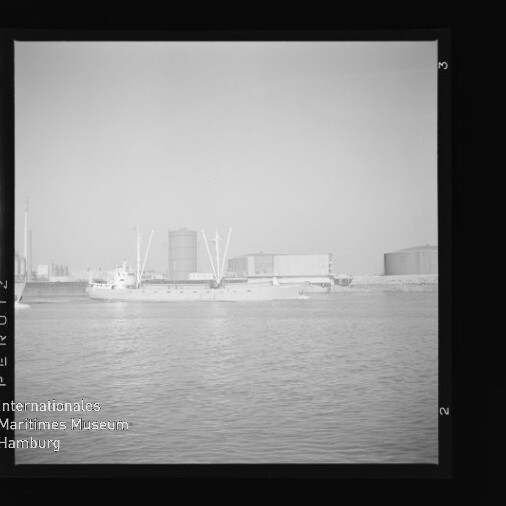 Schwarzweißaufnahme eines Frachtschiffs vor einem Industriehafen mit Tanks und Kränen, Wasser im Vordergrund.