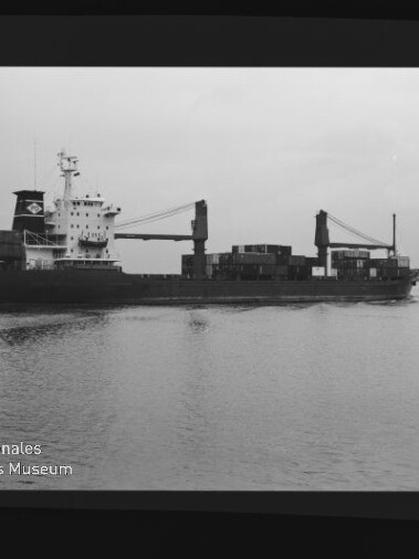 Frachtschiff mit Deckskranen und gestapelten Containern fährt seitlich auf ruhiger See, Museumslogo unten links.