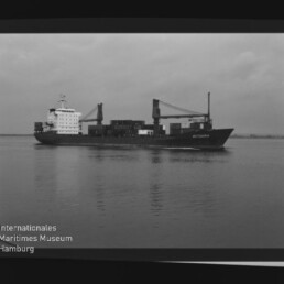 Schwarzweißaufnahme eines Frachtschiffs mit Deckskranen und Containern auf ruhigem Meer, Filmrand mit AGFA PAN 100 und Museumswasserzeichen.