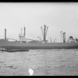 Frachtschiff mit mehreren Ladekränen und Masten, kleines Begleitboot im Vordergrund und Hafen im Hintergrund, Schwarzweißfoto