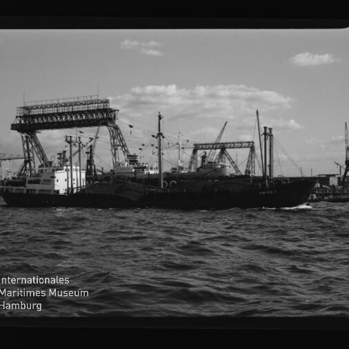 Frachtschiff auf bewegter See vor einem Hafen mit großen Portalkranen und Kränen; Museumssignet 