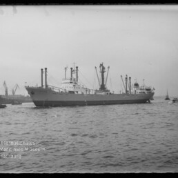 Frachtschiff mit mehreren Lademasten und Schleppern vor Hafenkränen, Wasser im Vordergrund.