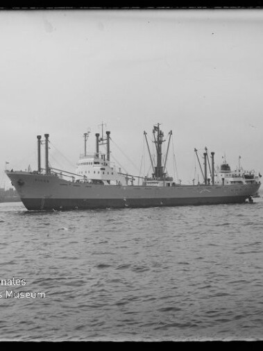 Frachtschiff mit mehreren Lademasten und Schleppern vor Hafenkränen, Wasser im Vordergrund.