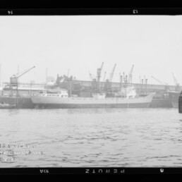Schiff am Industriehafen an der Kaimauer, mehrere Hafenkräne im Hintergrund und Wasser im Vordergrund.