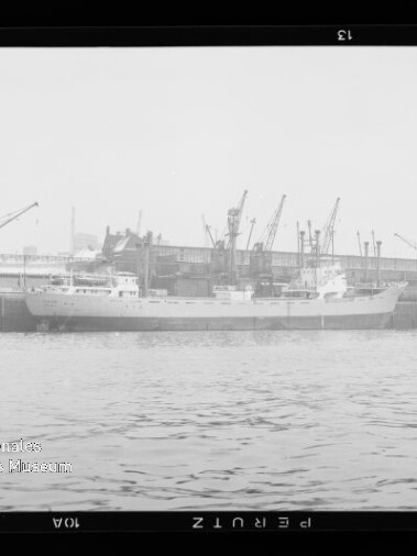 Schiff am Industriehafen an der Kaimauer, mehrere Hafenkräne im Hintergrund und Wasser im Vordergrund.