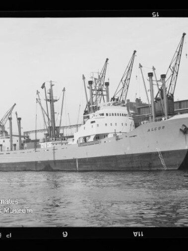 Frachtschiff ALCOR liegt am Kai vor Lagerhäusern, mehrere Hafenkräne darüber und ruhiges Wasser im Vordergrund.