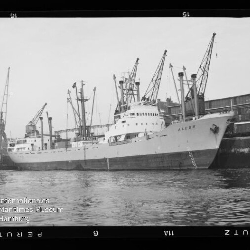 Frachtschiff ALCOR liegt am Kai vor Lagerhäusern, mehrere Hafenkräne darüber und ruhiges Wasser im Vordergrund.