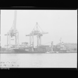 Hafen mit zwei großen Containerkranen, angelegtem Frachtschiff und Schlepper vor Lagerhallen; Museumsstempel links unten.