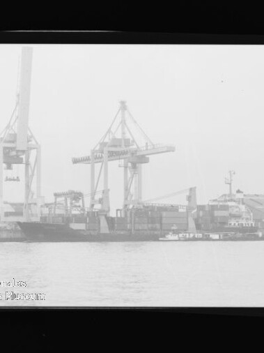 Hafen mit zwei großen Containerkranen, angelegtem Frachtschiff und Schlepper vor Lagerhallen; Museumsstempel links unten.