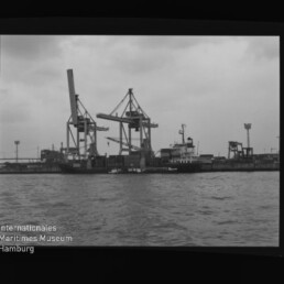 Hafen mit großen Containerkränen, angedocktem Frachtschiff, Wasser vorne und Wasserzeichen des Maritimen Museums Hamburg.