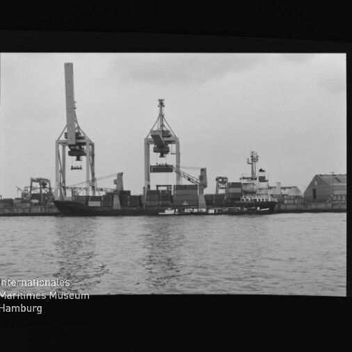Frachtschiff am Kai unter zwei großen Ladekränen, kleiner Schlepper im Vordergrund; Museumssignet links unten.